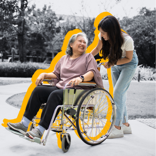 Younger woman walking with elderly woman in a wheelchair down a path on a bright day, smiling at each other